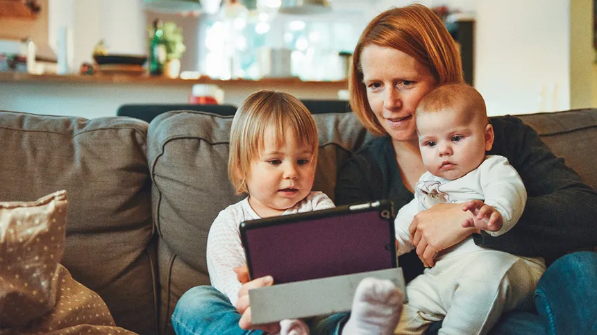 Mutter sitzt mit ihren zwei Kinder auf einem Sofa und zeigt ihnen was auf dem iPad