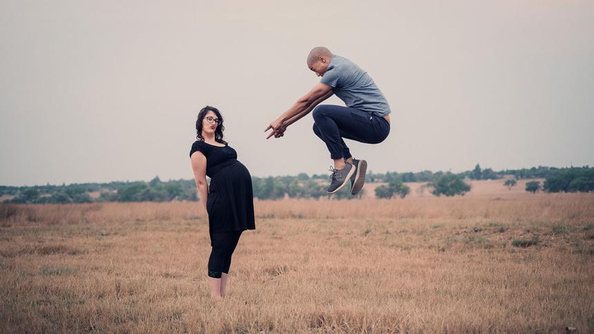 Frau in der 30. SSW steht mit ihrem Babybauch auf einem Feld, Mann springt in die Luft