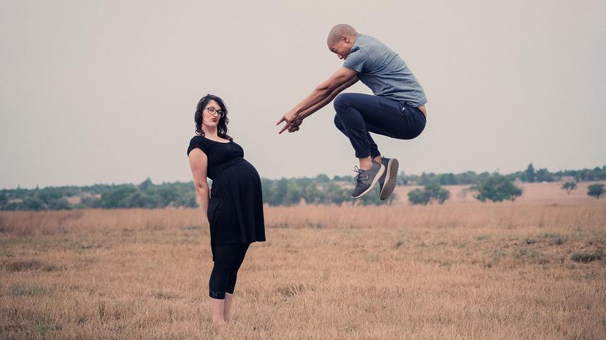 Schwangere Frau steht auf einem Feld. Partner zeigt auf Babybach