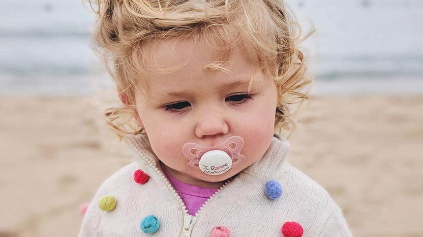 Blondes Kind mit Schnuller im Mund steht am Strand
