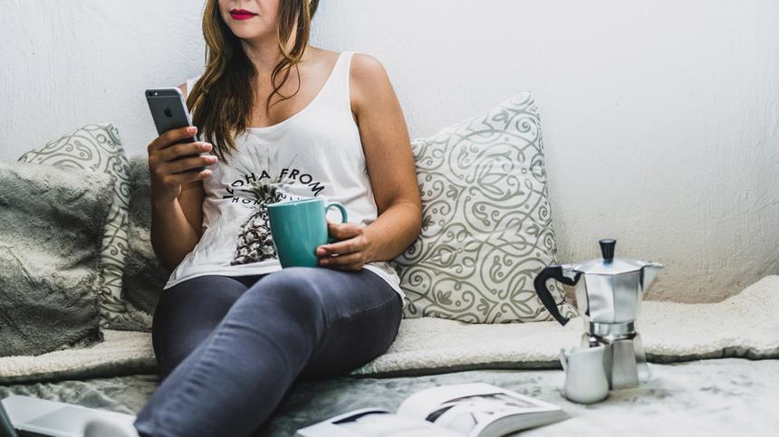 Eine Frau sitzt mit Handy und Kaffeetasse auf der Couch