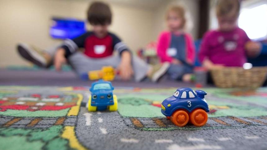 Zwei Spielzeugautos auf einem Spielteppich. Im Hintergrund sitzen drei Kleinkinder